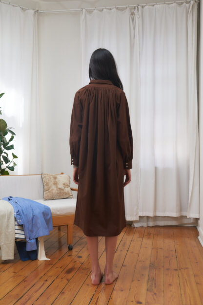 Brown shirt dress with gathered volume on the body. Long sleeves and mid-calf length. org. cotton poplin. this image shows back view.