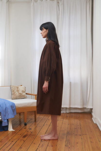 Brown shirt dress with gathered volume on the body. Long sleeves and mid-calf length. org. cotton poplin. this image shows side view.